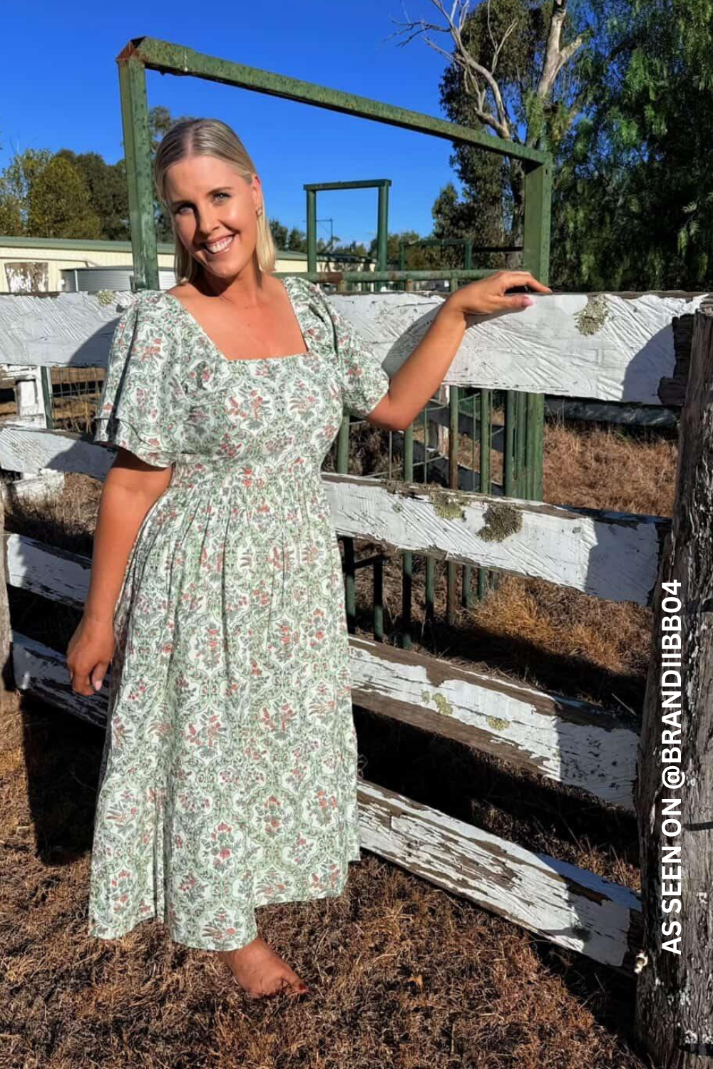 Woman in a floral dress standing next to a wooden fence with trees and buildings in the background.