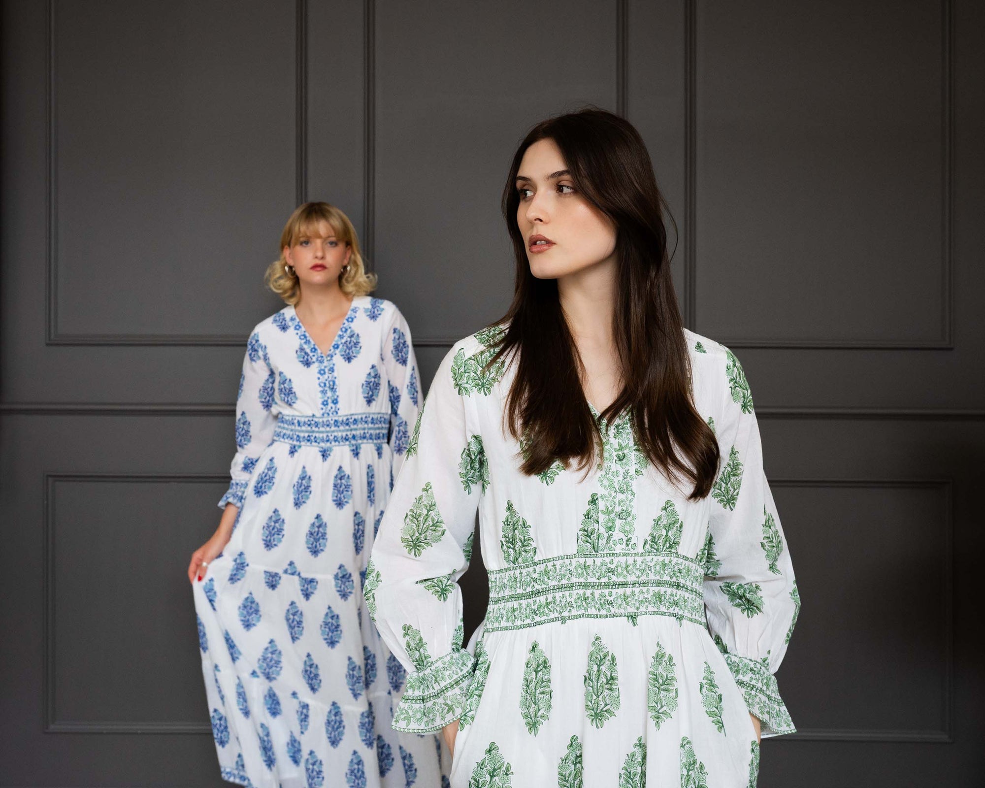 Two women wearing floral dresses against a dark paneled wall.