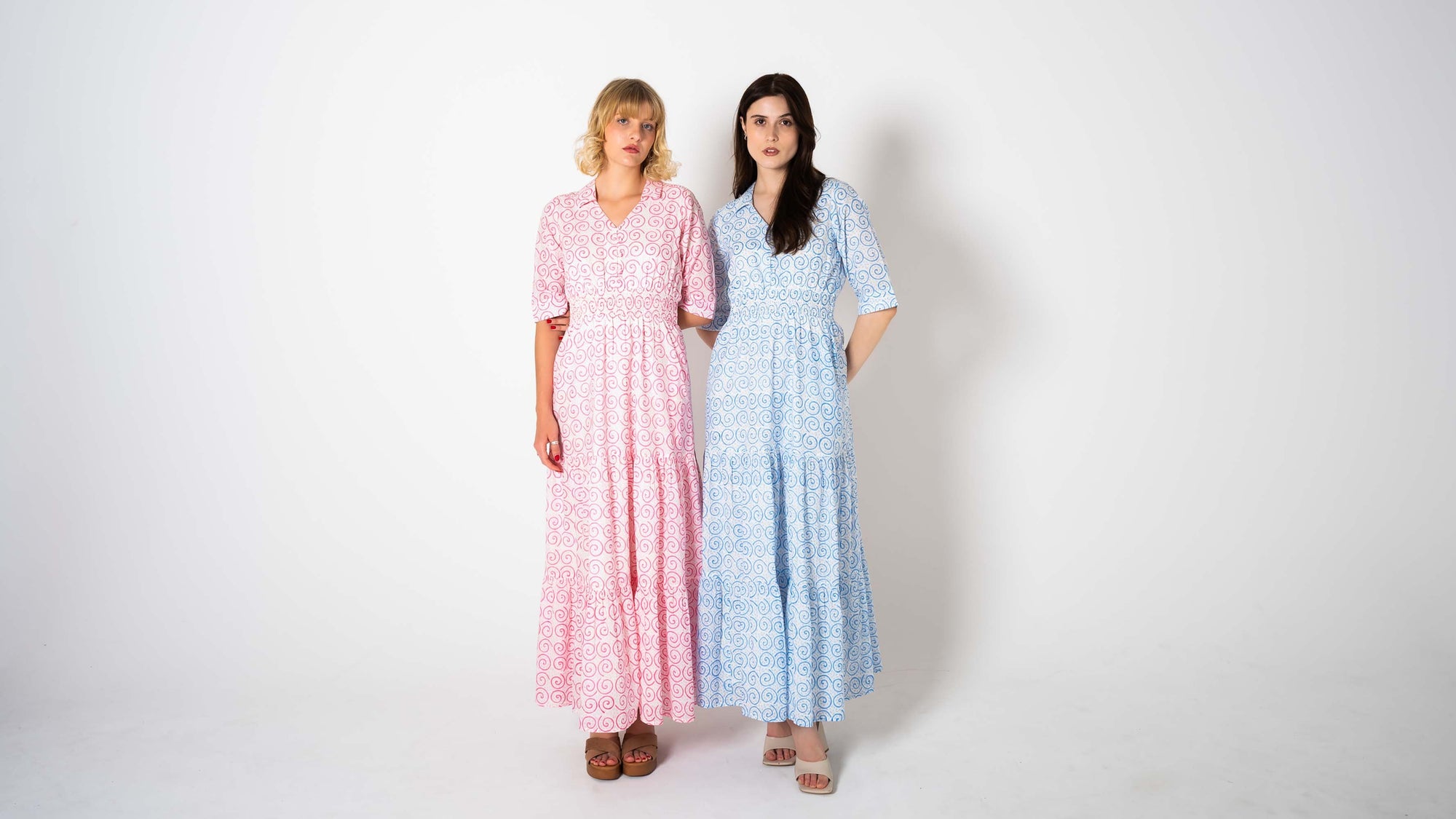 Two women wearing long, patterned dresses standing side by side against a white background.