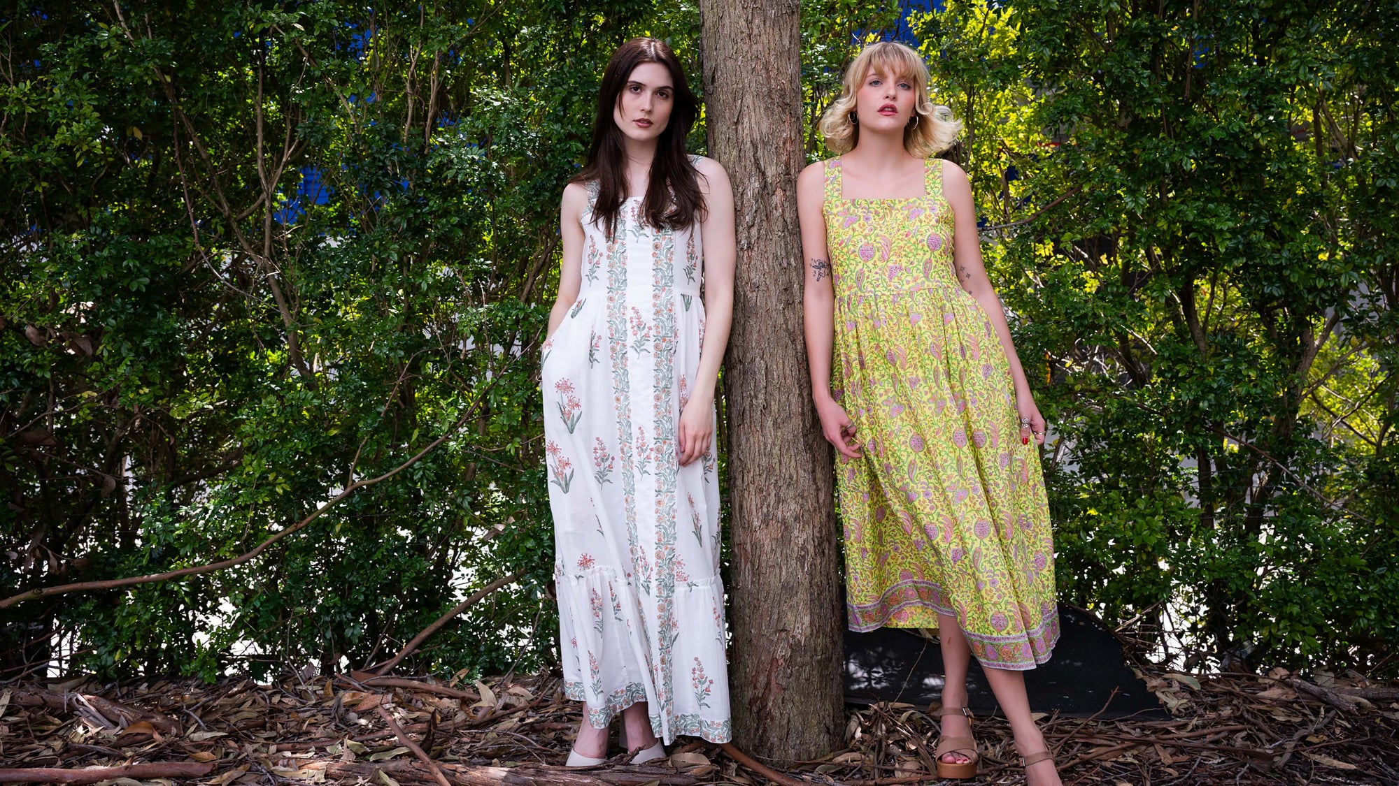 Two women standing outdoors, one in a white dress and the other in a green dress, against a natural background.